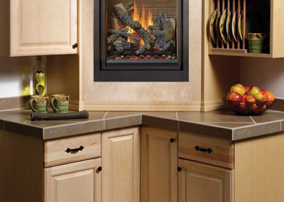 Stock photo of ProBuilder 24 CF Deluxe fireplace between kitchen cabinets sitting on the counter top with apples on the right and cups on the left.