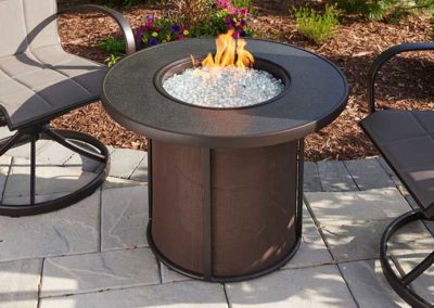 Stock photo of 32 Stonefire outdoor fire table with clear fire glass chairs on each side and landscaping in the background.