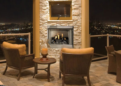 Stock photo of 36" Gas Outdoor Fireplace with two outdoor chairs and a tv with a beautiful view of a lighted city.