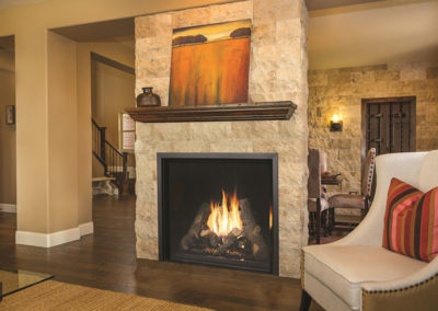 Stock photo of CF fireplace insert with large flames with stone surround. There is a picture of trees on the wooden mantel and a chair to the right. The background is a dining room.