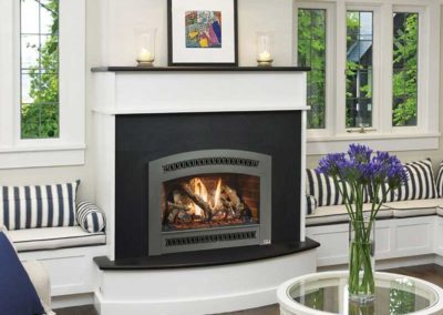 Stock photo of TRV 25K fireplace insert with black surround and white mantel with windows on each side and window seats below. Beautiful purple flowers on coffee table.