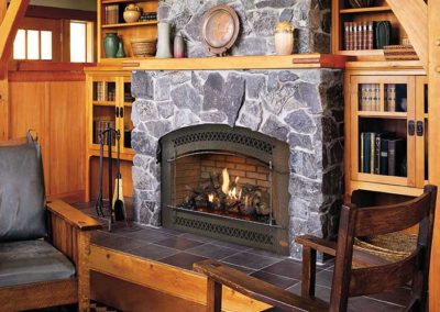 Stock photo of TRV 31k fireplace gas fireplace insert with stone surround and tile hearth. The room is wood with bookcases and shelves and a chair on each side of the fireplace.