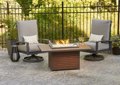 Stock photo of Kenwood Fire Table on concrete patio with two gray out rocking chairs black fencing and landscaping in the background.