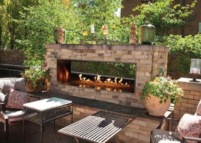 Stock photo of uutdoor Linear See Through Gas Fireplace with chairs and two coffee tables - two potted plants beside fireplace and trees in the background.