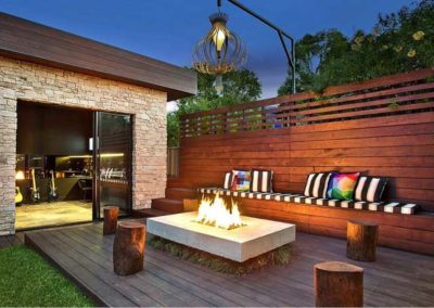 Rectangular fire pit on back patio with beautiful stained privacy fence in the background and tree stumps to sit around firepit and seating along fence.