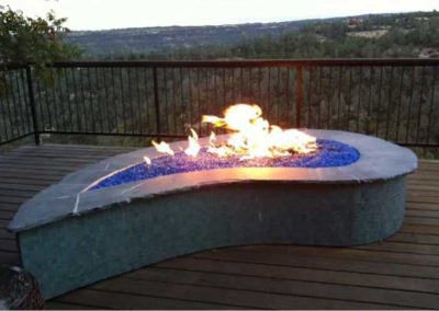 Stock photo of tear drop shaped fire pit with blue fire glass on a wooden deck overlooking trees.