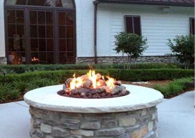 Stock photo of round fire pit on patio. Home in the background has a large beautiful arched window. The home is white.