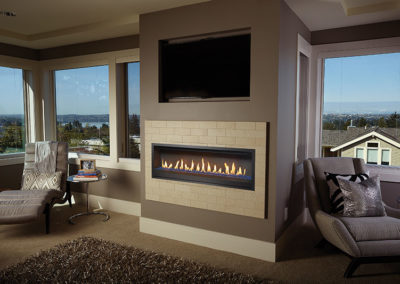 Stock photo of 54 fireplace gas insert with tv above and windows all around the room. There is a lounge chair on each side and a lake and home looking out of the window.