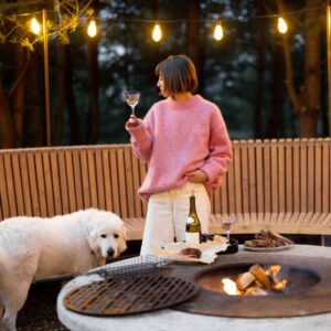 a woman in a pink sweater with a glass of wine standing next to a dog by a fire pit